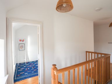 A hallway with wooden railing and a doorway showing a room with a blue rug and framed pictures at Ynys Faelog Cottage in Ynys Faelog near Menai Bridge