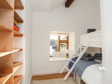 A bedroom with a white bunk bed blue blankets wooden shelves and a window with a lamp at Ynys Faelog Cottage in Ynys Faelog near Menai Bridge