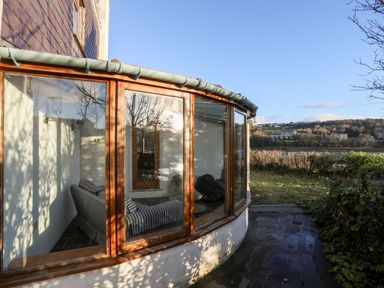 A curved glass window showing a room with a striped sofa and a stone pathway leading to a green lawn outside at Ynys Faelog Cottage in Ynys Faelog near Menai Bridge