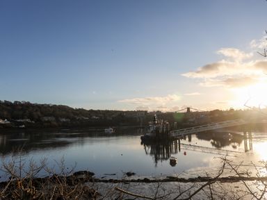 A river with a dock and boats and a suspension bridge in the background at Ynys Faelog Cottage in Ynys Faelog near Menai Bridge