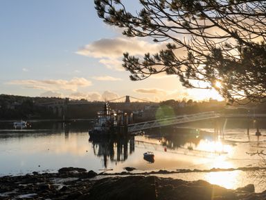 A river view with a boat docked near a pier and a suspension bridge in the background at Ynys Faelog Cottage in Ynys Faelog near Menai Bridge