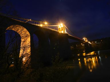 A large stone bridge with arches and suspension cables lit up at night over a river at Ynys Faelog near Menai Bridge