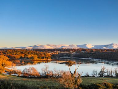 A river with a suspension bridge in the background and snow-covered mountains at Ynys Faelog near Menai Bridge
