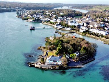 Aerial view of a coastal area with an island featuring houses trees and a tennis court near a town with bridges in the background at Ynys Faelog near Menai Bridge
