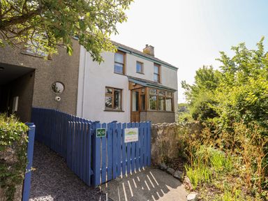 A white two-story house with wooden windows and blue gate surrounded by plants at Ynys Faelog Cottage Ynys Faelog near Menai Bridge