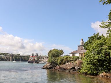 A house surrounded by trees on a rocky shoreline with boats and a suspension bridge in the background at Ynys Faelog near Menai Bridge