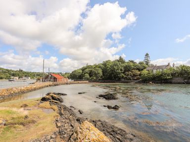 A coastal scene with rocky shore a red boathouse trees and buildings at Ynys Faelog Cottage in Ynys Faelog near Menai Bridge