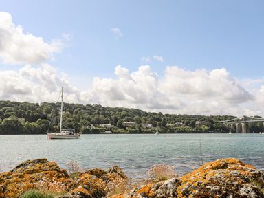 A body of water with a sailboat near grassy hills and a bridge in the background at Ynys Faelog Cottage in Ynys Faelog near Menai Bridge