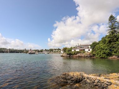 A waterfront house surrounded by trees near a body of water with a bridge and boats in the background at Ynys Faelog Cottage in Ynys Faelog near Menai Bridge