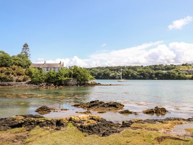 A coastal scene with a house, trees, rocks in the water, and a sailboat at Ynys Faelog Cottage in Ynys Faelog near Menai Bridge
