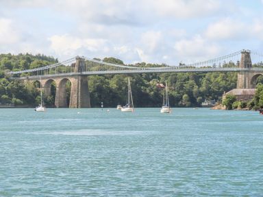 A suspension bridge over a river with sailboats and trees in the background at Ynys Faelog near Menai Bridge