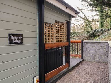 An outdoor area with a grey wooden wall and a sign reading Buttermilk Bridge next to a gravel path and wooden railing at Buttermilk Bridge in Dungiven