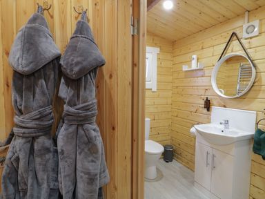 A bathroom with wooden walls a white sink a round mirror a toilet and two gray bathrobes hanging on hooks at Buttermilk Bridge in Dungiven