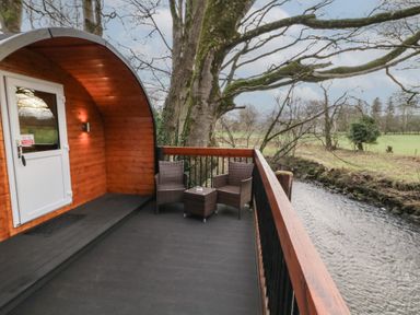 A wooden cabin balcony with two wicker chairs and a small table overlooking a river in a rural area at Pine in Dungiven Londonderry