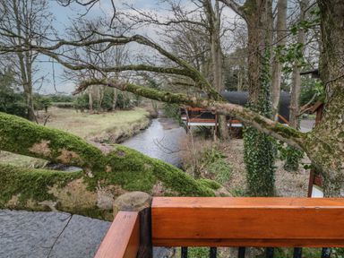 A wooden deck overlooking a moss-covered tree and a narrow river with houses and trees along the bank at Pine in Dungiven Londonderry