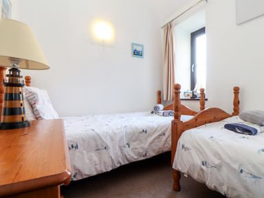 A bedroom with two single wooden beds a small wooden table with a lighthouse lamp and a window with beige curtains at The Old Piggeries in Gulval near Penzance