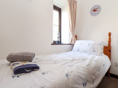 A small bedroom with a single bed with nautical themed bedding a small lighthouse decoration on the windowsill and a wall clock at The Old Piggeries in Gulval near Penzance