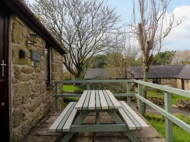 A stone building with a wooden picnic table on a patio and trees in the background at The Old Piggeries in Gulval near Penzance