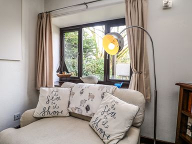 A beige armchair with two cushions and a sheep-patterned blanket near a window with beige curtains and a standing lamp at The Old Piggeries in Gulval near Penzance