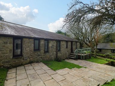 A stone building with multiple windows and a small porch with steps surrounded by grass and trees at The Old Piggeries in Gulval near Penzance