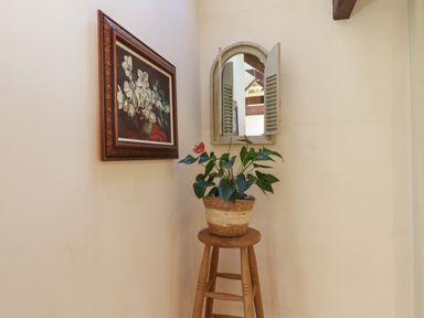 A corner with a wooden stool holding a potted plant below a framed flower painting and a shuttered mirror at The Calf Pen in Colyton