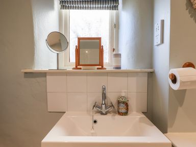 A bathroom sink with a faucet and soap bottle below a window with two mirrors on the windowsill and a toilet paper roll on the wall at The Calf Pen in Colyton