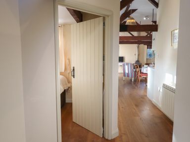 A hallway with a partially open door leading to a bedroom and a view of a living area with wooden beams and striped chair at The Calf Pen in Colyton