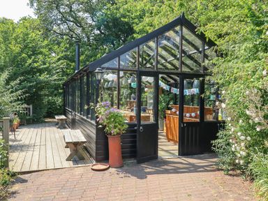 A greenhouse with open doors decorated for a birthday with benches outside surrounded by greenery at The Calf Pen in Colyton
