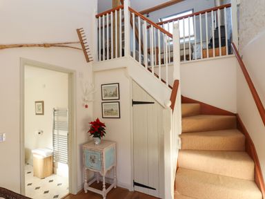 A staircase with beige carpet and wooden handrails next to a small blue cabinet with a red plant and two framed pictures on the wall at The Hay Loft in Colyton