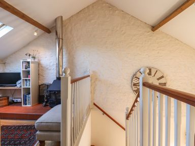 An attic room with a wood stove a beige ottoman a bookshelf a television and a large wall clock at The Hay Loft in Colyton