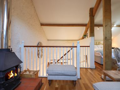 An upper floor with a wood burning stove a basket with firewood a gray ottoman wooden beams a bed and a wall clock at The Hay Loft in Colyton