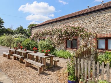 An outdoor seating area with wooden benches and tables next to a stone building with flowering plants at The Hay Loft in Colyton