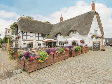 An outdoor seating area with picnic tables and umbrellas outside a thatched roof pub with flower boxes on a cobblestone surface at The Old Bakehouse in Avebury