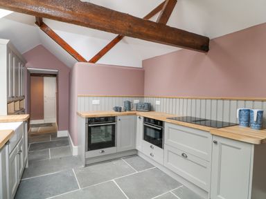 A kitchen with grey cabinets wooden countertops a stove ovens mugs and kettle under wooden beams at The Old Bakehouse in Avebury