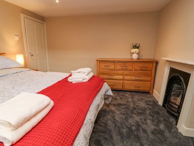 A bedroom with a bed covered with gray patterned bedding and a red throw a wooden dresser with a vase of flowers and a fireplace at The Old Bakehouse in Avebury