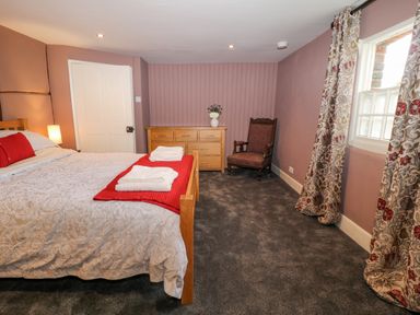 A bedroom with a bed with white and red bedding a wooden dresser an armchair and floral curtains at The Old Bakehouse in Avebury