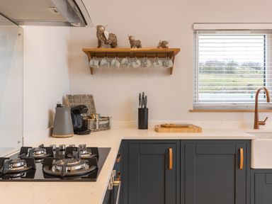 A kitchen with a gas stove a knife block a sink a window with blinds and a wooden shelf with hanging mugs and decorative figures at Stags Mount in Thornton Dale