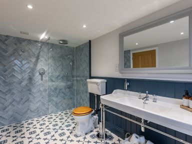 A bathroom with a shower area with blue tiled walls a toilet with wooden seat a double sink with a large mirror and patterned floor tiles at Stags Mount in Thornton Dale