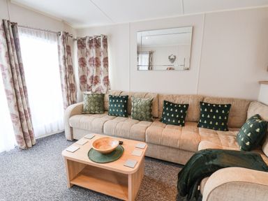 A living room with a beige sectional sofa adorned with green and patterned cushions a wooden coffee table with a bowl and coasters curtains with tree patterns and a wall mirror at Forest Star in Cayton