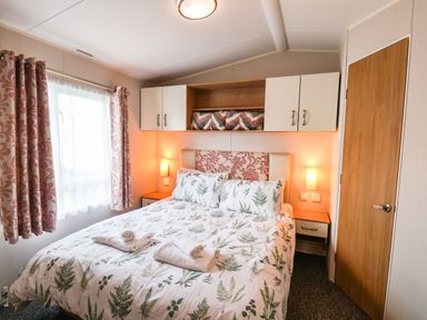 A bedroom with a bed with patterned bedding and towels on it two bedside tables with lamps a window with patterned curtains a wooden door and overhead cabinets at Forest Star in Cayton