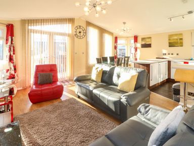 An open living area with gray sofas, a red chair, dining table with chairs, kitchen island, and tall windows at Dream Catcher Lodge in Carnforth
