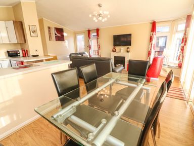A living and dining area with a glass dining table and black chairs a kitchen counter with appliances sofas a wall-mounted TV and floral curtains at Dream Catcher Lodge in Carnforth