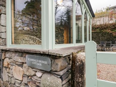 Stone wall with a sign reading Scandale Bridge Cottage next to a window with green frames and an outdoor patio with chairs at Scandale Bridge Cottage Ambleside