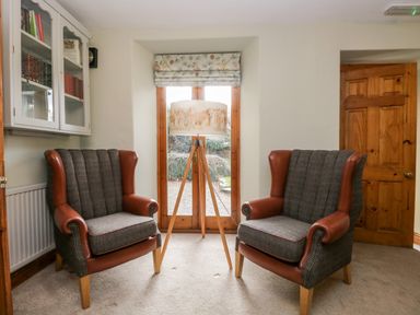 Two armchairs facing each other with a tripod floor lamp between them in a room with wooden door and glass cabinet at Scandale Bridge Cottage Ambleside