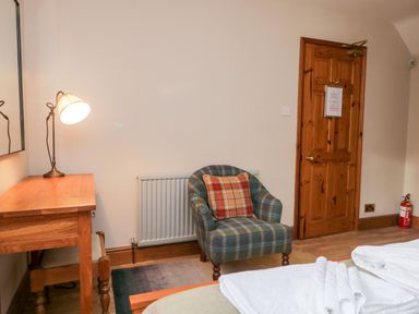 A room with a wooden desk and lamp a patterned armchair with a cushion a wooden door and folded towels on a bed at Scandale Bridge Cottage in Ambleside