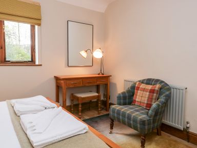 A bedroom with a wooden desk and stool a mirror a table lamp a plaid armchair and a bed with folded towels at Scandale Bridge Cottage in Ambleside
