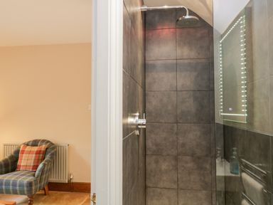 A tiled shower area with a rain shower head and an LED-lit mirror inside a bathroom next to a room with a checkered armchair and a radiator at Scandale Bridge Cottage in Ambleside