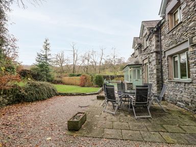 An outdoor patio area with a glass table and six chairs next to a stone house with green doors and windows a garden with gravel and grass at Scandale Bridge Cottage in Ambleside