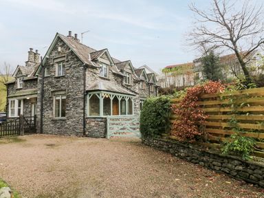A stone cottage with multiple windows and a gravel driveway next to a wooden fence and trees at Scandale Bridge Cottage in Ambleside