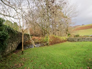 A grassy area with leafless trees and a stone bridge over a stream at Scandale Bridge Cottage in Ambleside
